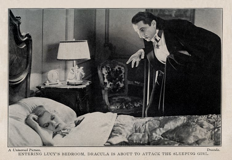 A black-and-white photo shows a woman lying in a bed sleeping, while a man wearing a black jacket and white shirt leers over her.