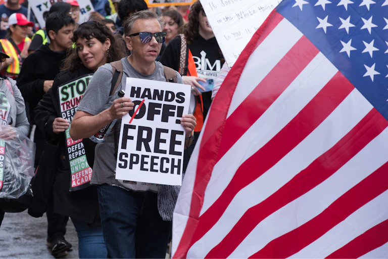 A group of protesters hold signs – with one reading 'Hands Off! Free Speech' – with an American flag in the foreground.
