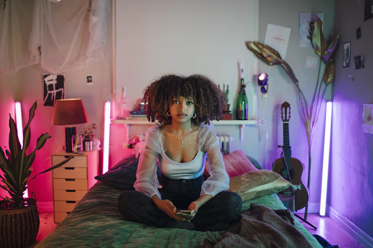 A young woman sits cross-legged on a twin bed in what appears to be a childhood bedroom.