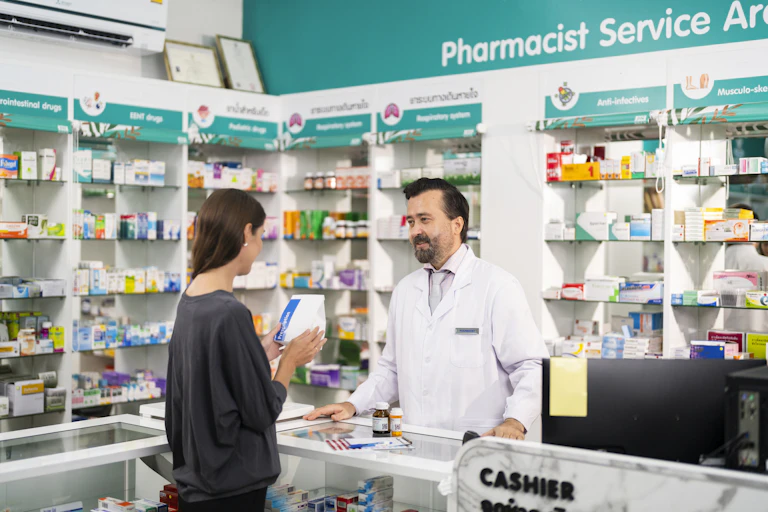 A bearded man in a white coat stands at a pharmacy counter talking with a young woman in a gray shirt.