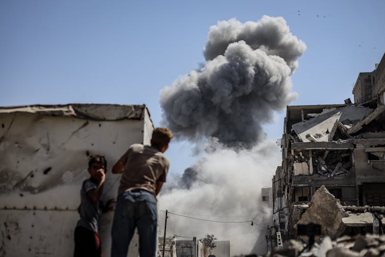 Two people look through a gap in the wall as a plume of smoke rises in the background.