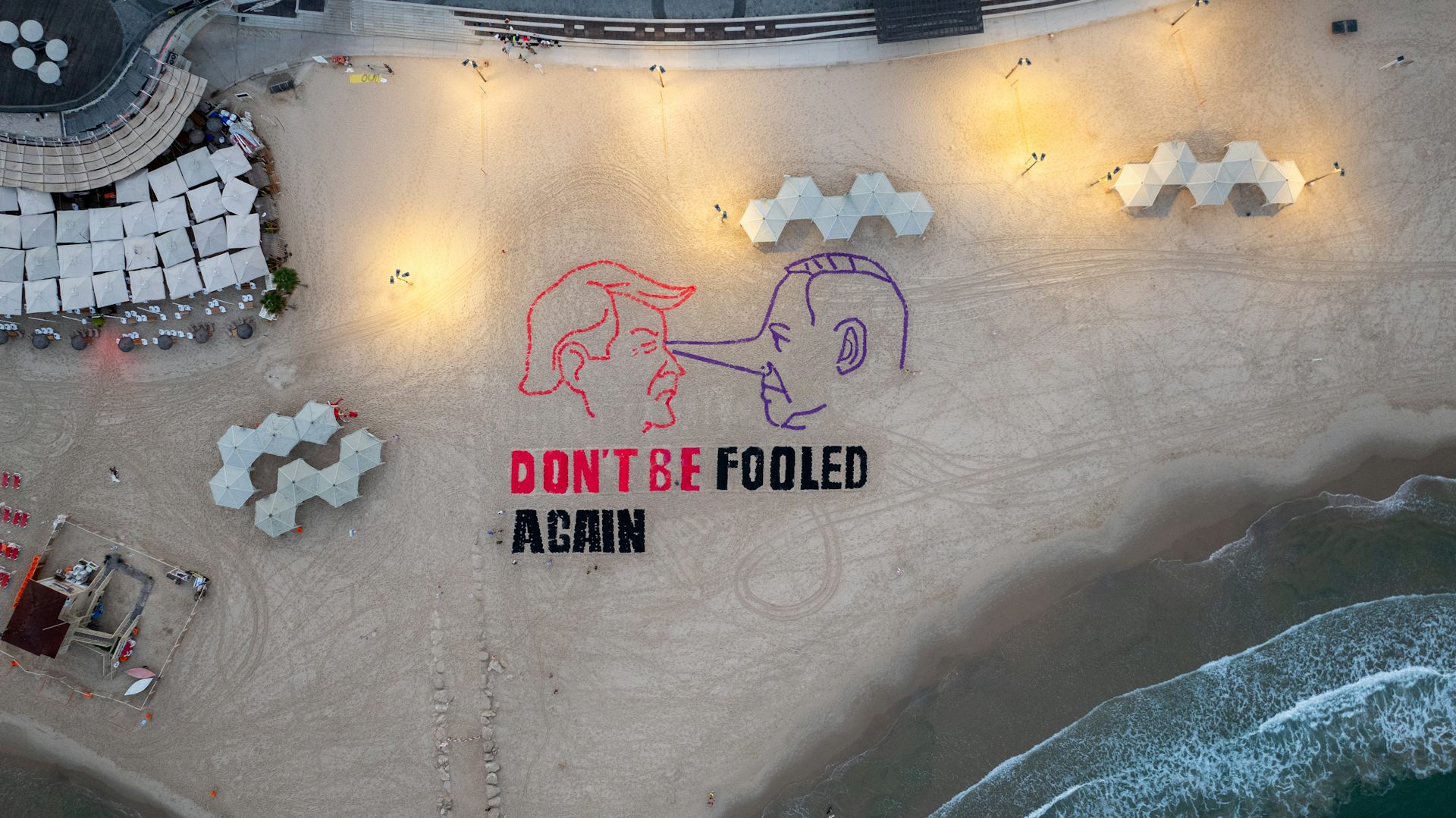 A display on a beach shows the heads of two men, one with an extended Pinocchio nose, indicating a liar. Underneath it says 'Don't be fooled again.'