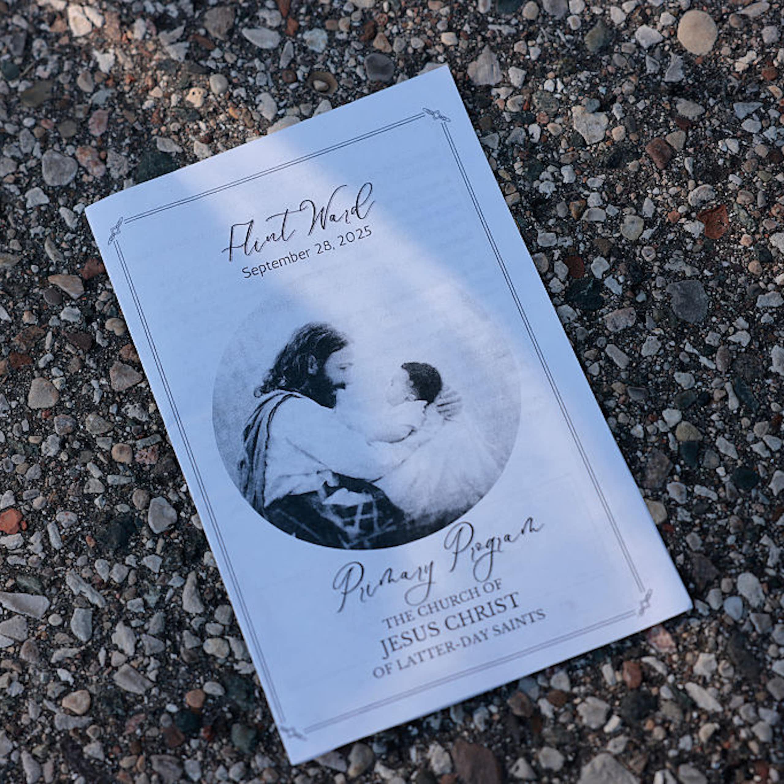 A white booklet with an illustration of Jesus embracing a child sits on an asphalt surface.