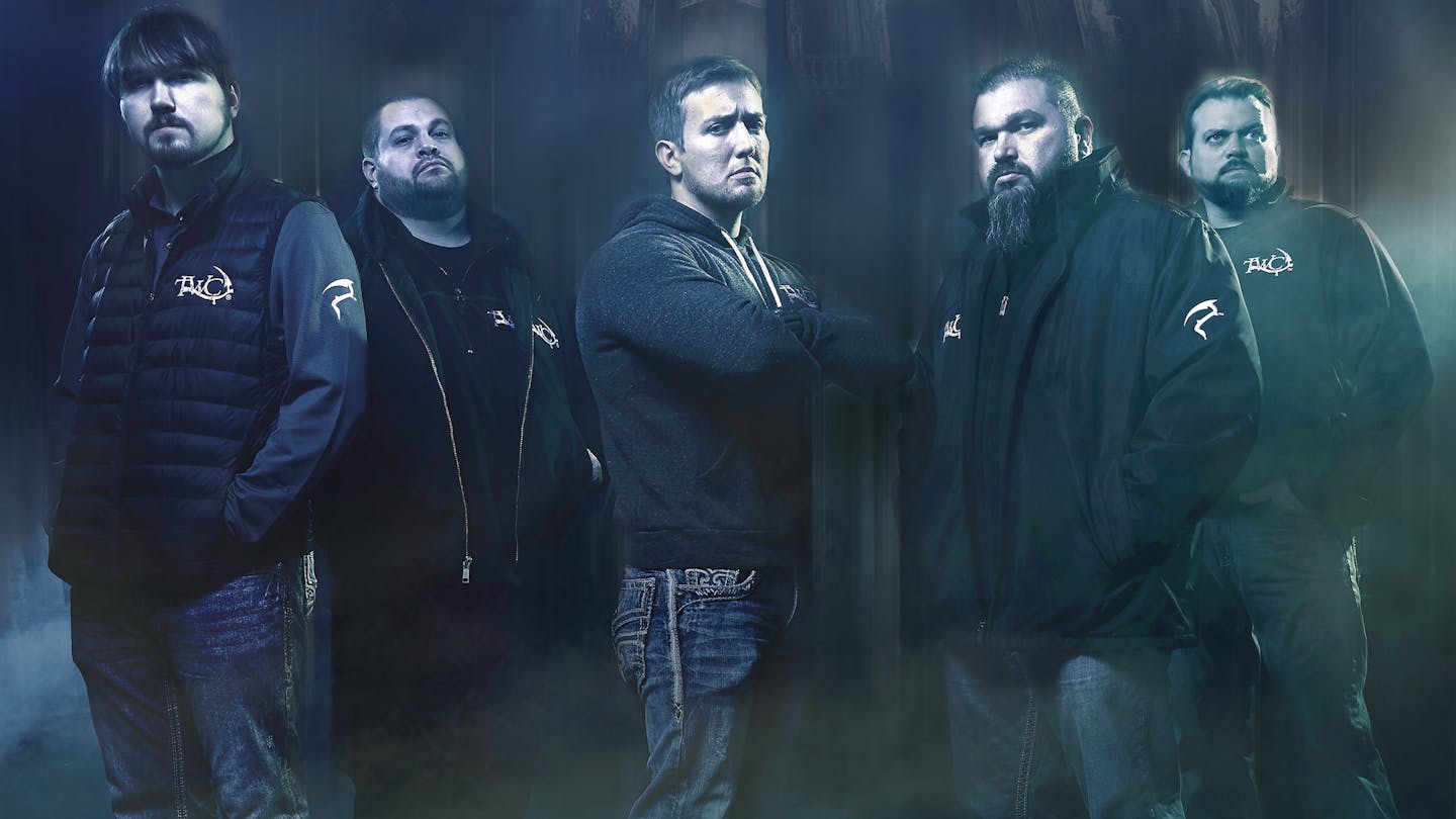 A group of burly, straight-faced men pose against a dark, shadowy bakground, in a promotional photo for a ghost-hunting TV show.