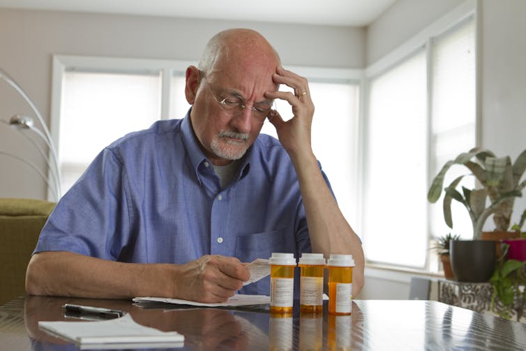 Um homem idoso olha para uma folha de papel com confusão. Ele tem três frascos de medicamentos prescritos na mesa à sua frente.