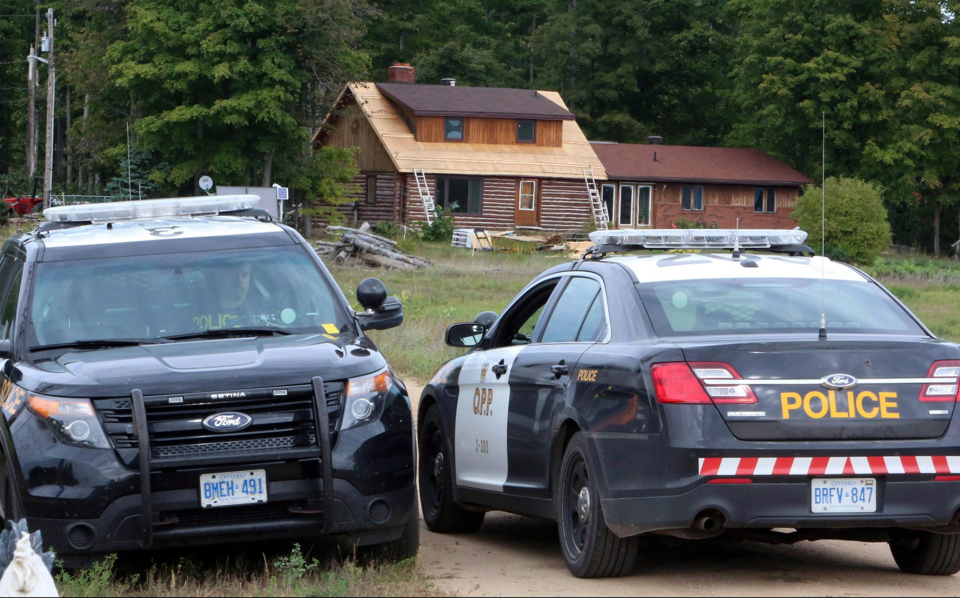Two police cruisers sit at the end of a driveway of a rural home.