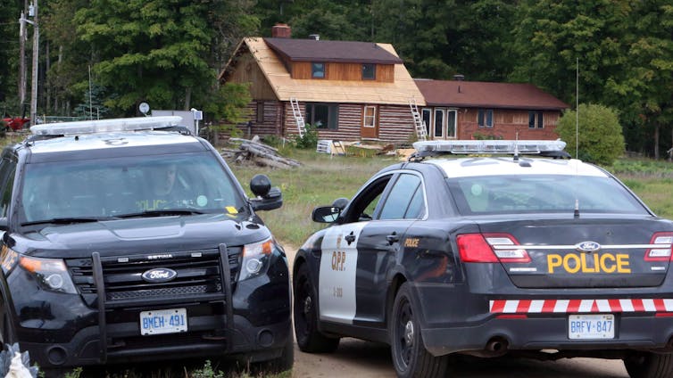 Two police cruisers sit at the end of a driveway of a rural home.