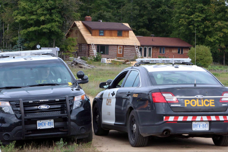 Two police cruisers sit at the end of a driveway of a rural home.