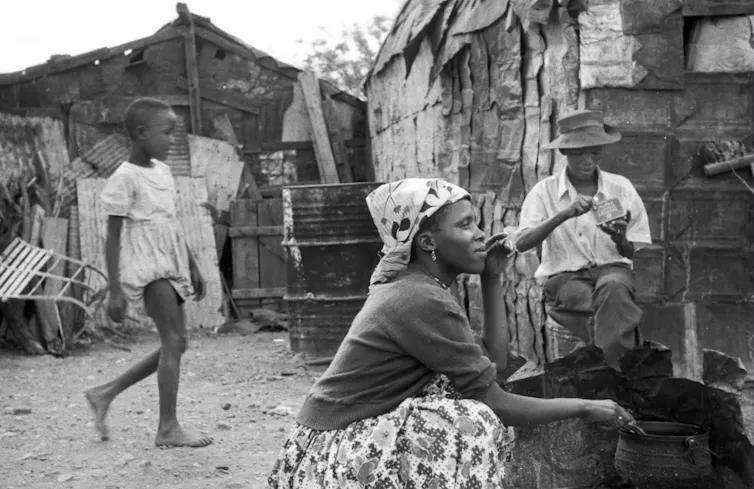 A family in a yard in front of a shack, a woman cooking, a man sitting and a child walking.