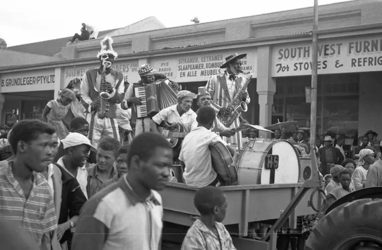 A black and white photo of a street lined with people and a cart moving down the road holding a band of minstrels in costume and make-up.