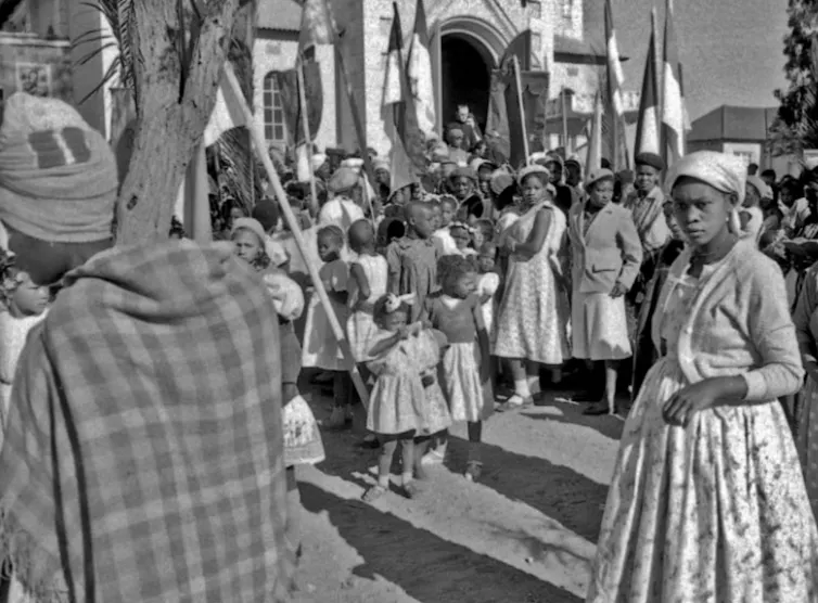 A black and white photo of mostly women in smart dresses and hats in front of a building.
