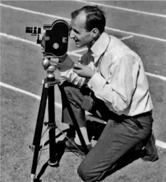 A black and white photo of a man in shirt and tie kneeling as he looks through a large old fashioned camera.
