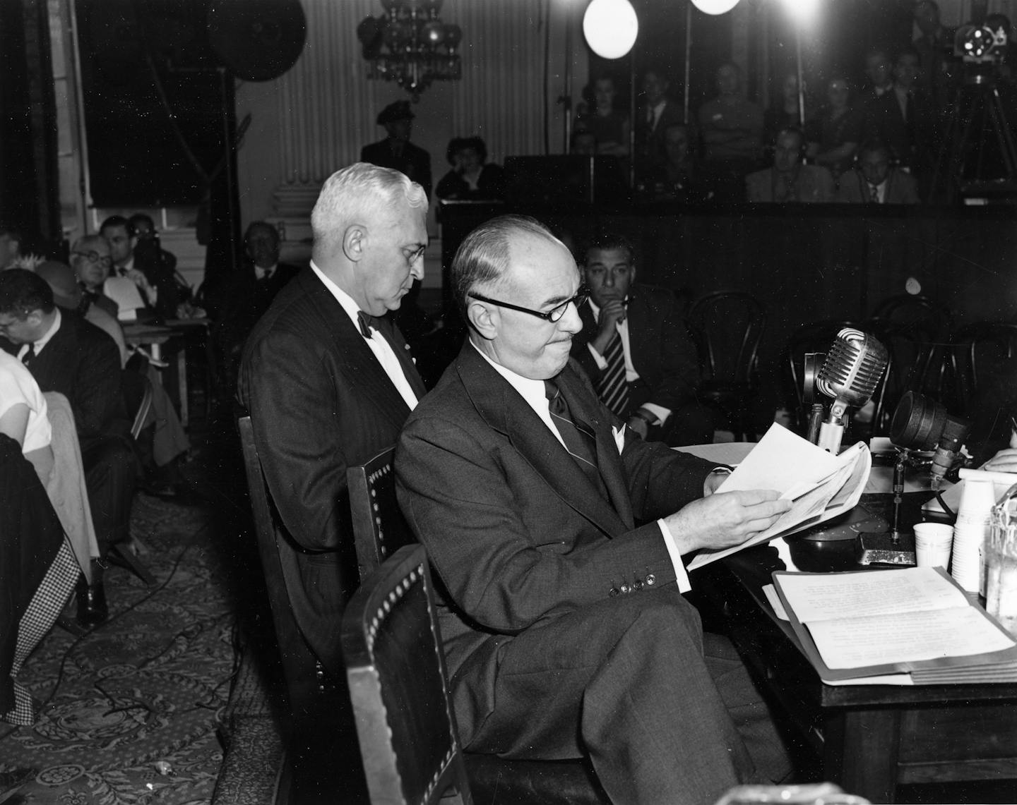 A black-and-white photo shows two men dressed in suits sitting in front of a desk.