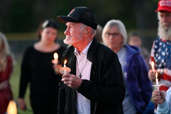 A man with a white beard, wearing a black sports cap and a cross necklace, somberly holds up a lit candle amid a crowd outside.