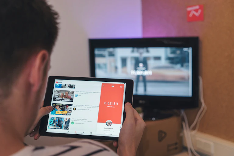 a young man watches a tablet showing YouTube with a computer screen in the background