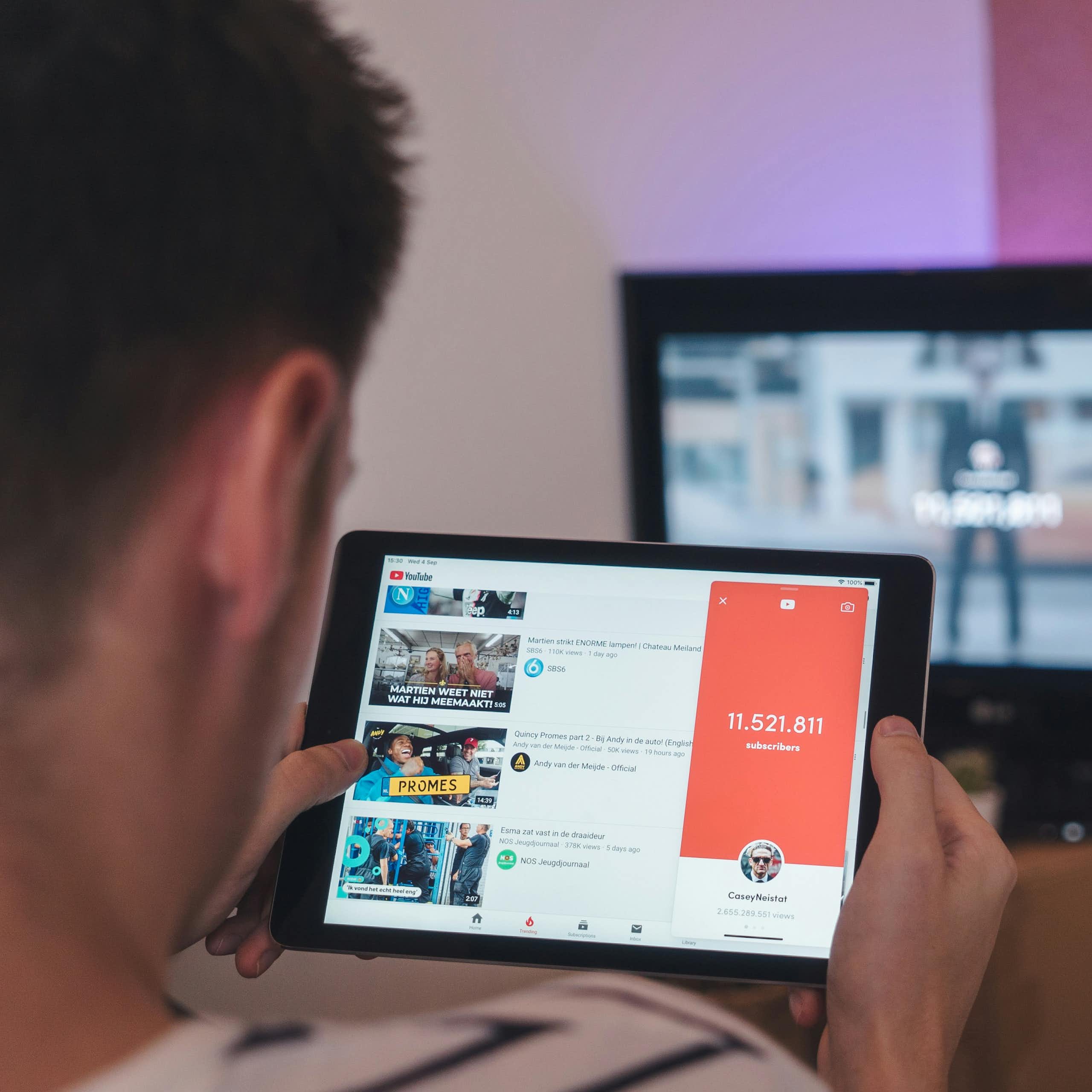 a young man watches a tablet showing YouTube with a computer screen in the background