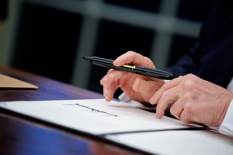 Two hands shown holding a black pen as they hover above white paper with a large signature on it.