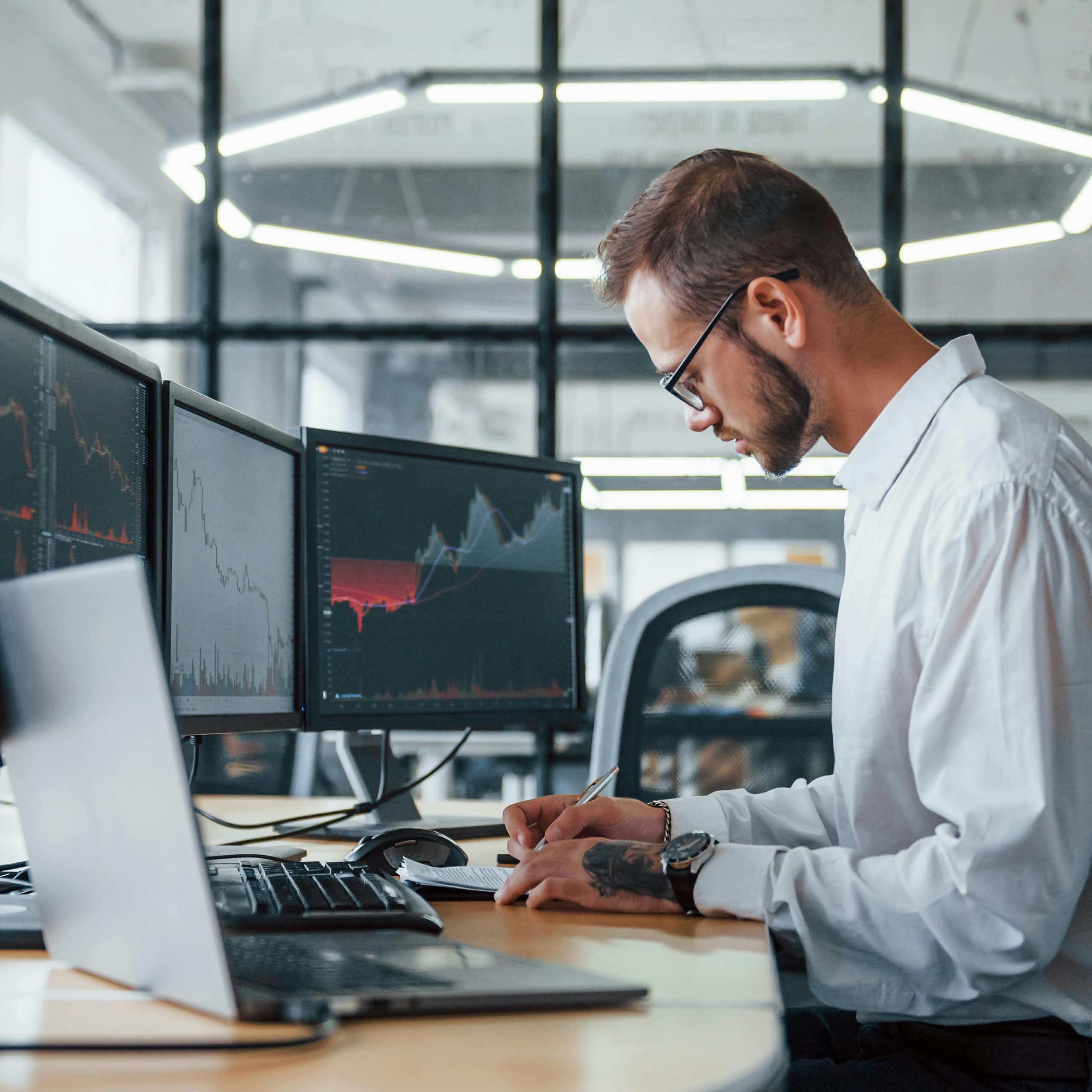 A person writes on a notepad while seated at a desk with three computer monitors and an open laptop on it