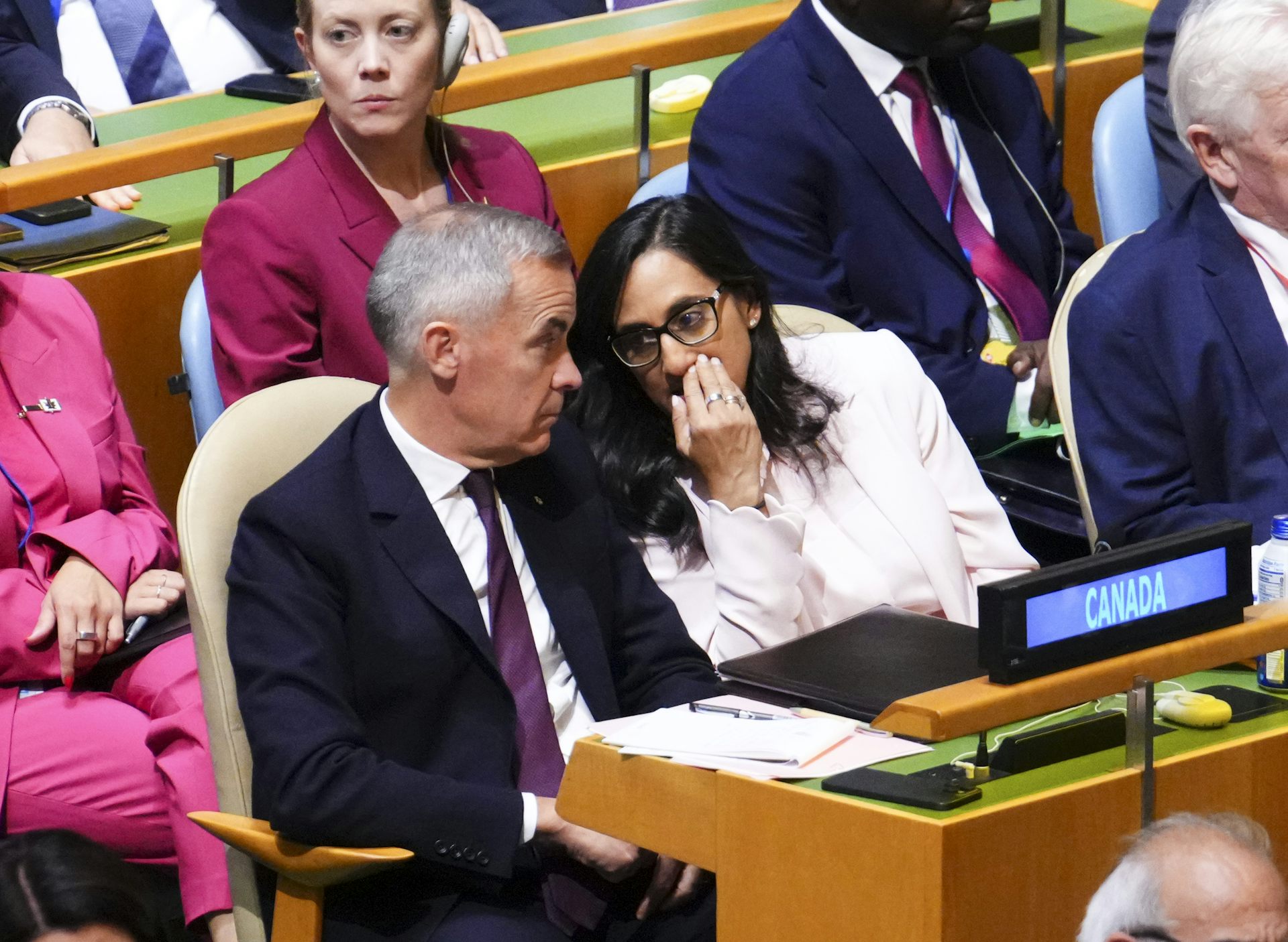A woman with dark hair and glasses speaks behind her hand to a man with short grey hair wearing a navy suit.