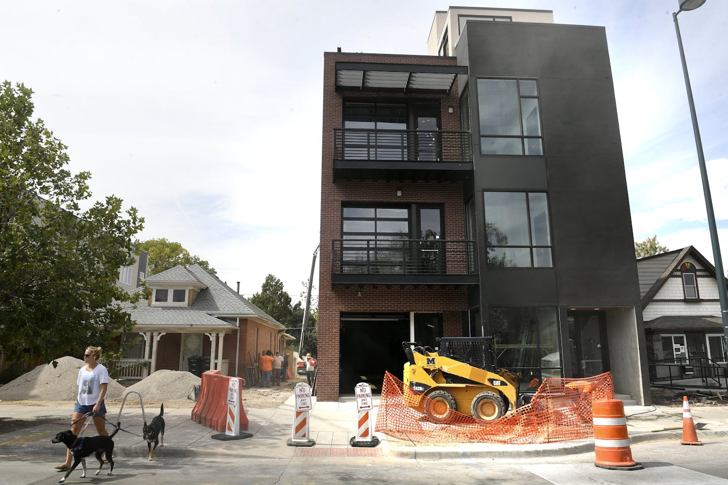 A woman walks two dogs near a gleaming new brown building that towers above neighboring homes. Orange traffic cones and a temporary fence are in the foreground.
