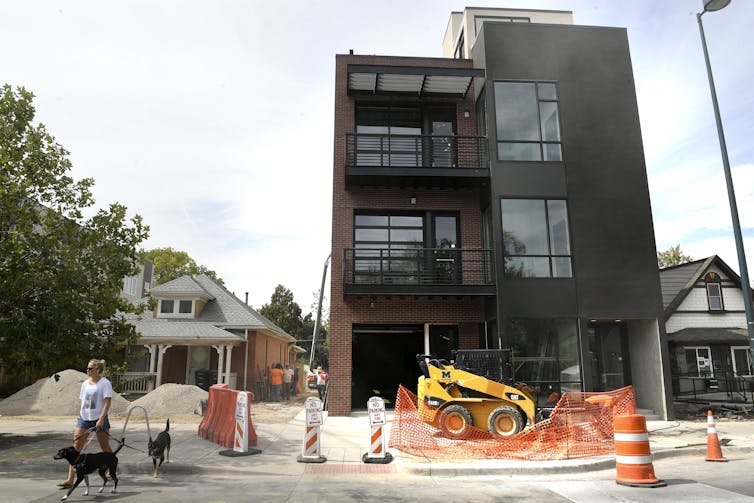 A woman walks two dogs near a gleaming new brown building that towers above neighboring homes. Orange traffic cones and a temporary fence are in the foreground.
