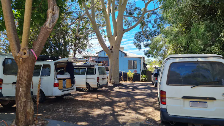 The exterior of a blue building back from a dirt parking area filled with white vans