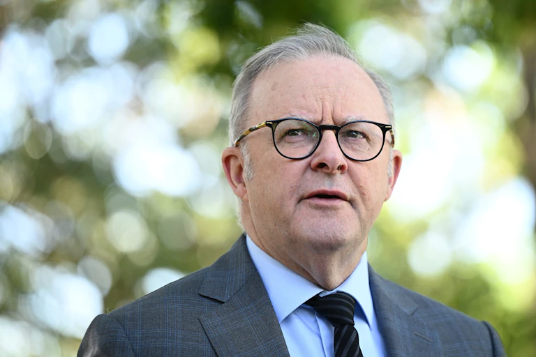 A grey haired man wearing glasses against backdrop of trees