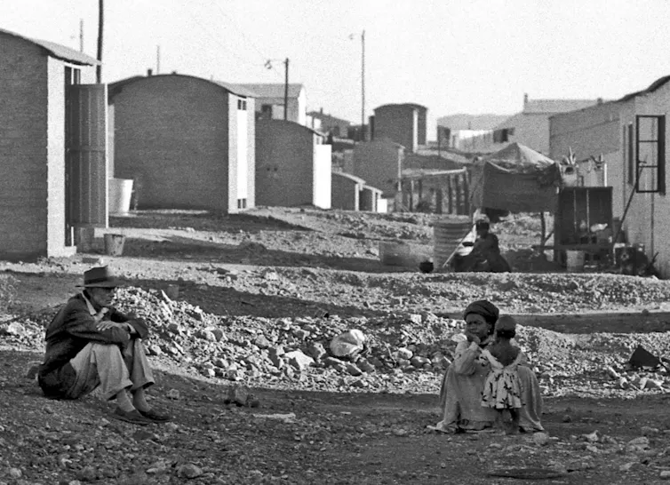 A forlorn couple sits on the ground amid building rubble. Behind them is a row of small new houses.