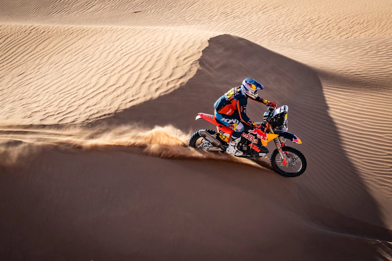 Un hombre conduce una moto naranja entre las dunas del desierto.