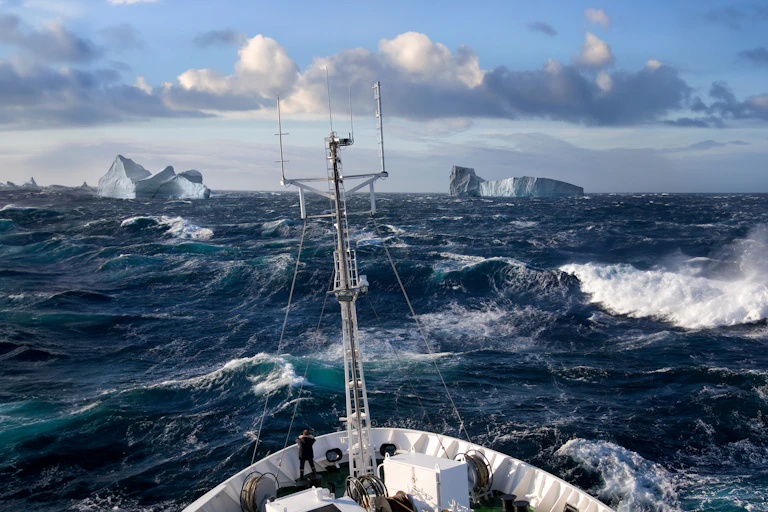 A rough sea with a ship's prow just visible with icebergs in the distance.