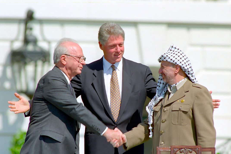 US president Bill Clinton holds his hands wide as Israeli prime minister Yitzhak Rabin and Palestinian leader Yasser Arafat shake hands.
