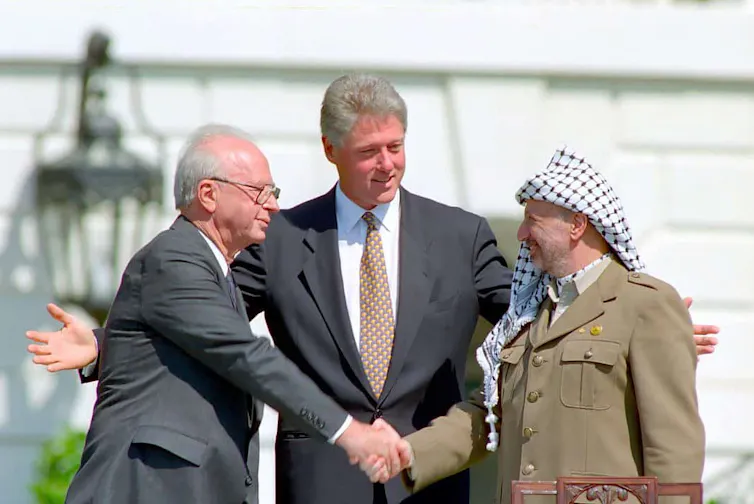 US president Bill Clinton holds his hands wide as Israeli prime minister Yitzhak Rabin and Palestinian leader Yasser Arafat shake hands.