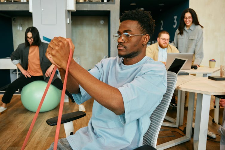 People in an office, one of whom sitting on a balance ball and using an exercise band to strengthen an arm