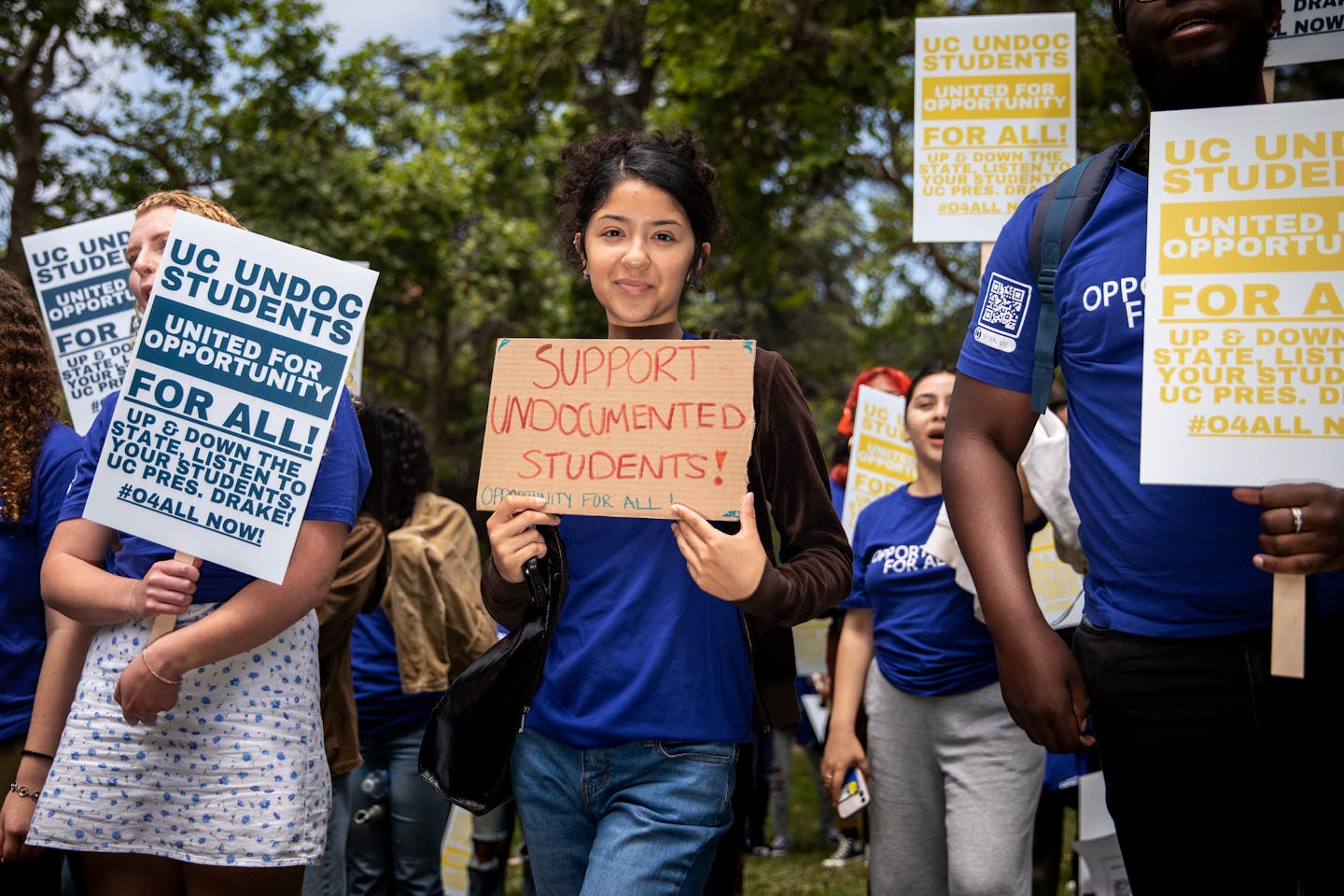 A young woman with brown hair in a blue shirt holds a sign that says 'Support Undocumented Students.' Other people stand around her in blue shirts and also hold signs.