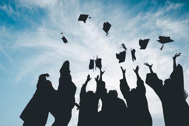 People at a graduation ceremony are seen in silhouette throwing their mortarboards skyward.