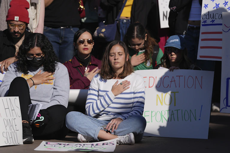A group of young people sit on the ground and close their eyes and hold their hands over their chests. One person has a sign that says 'Education not Deportation.'