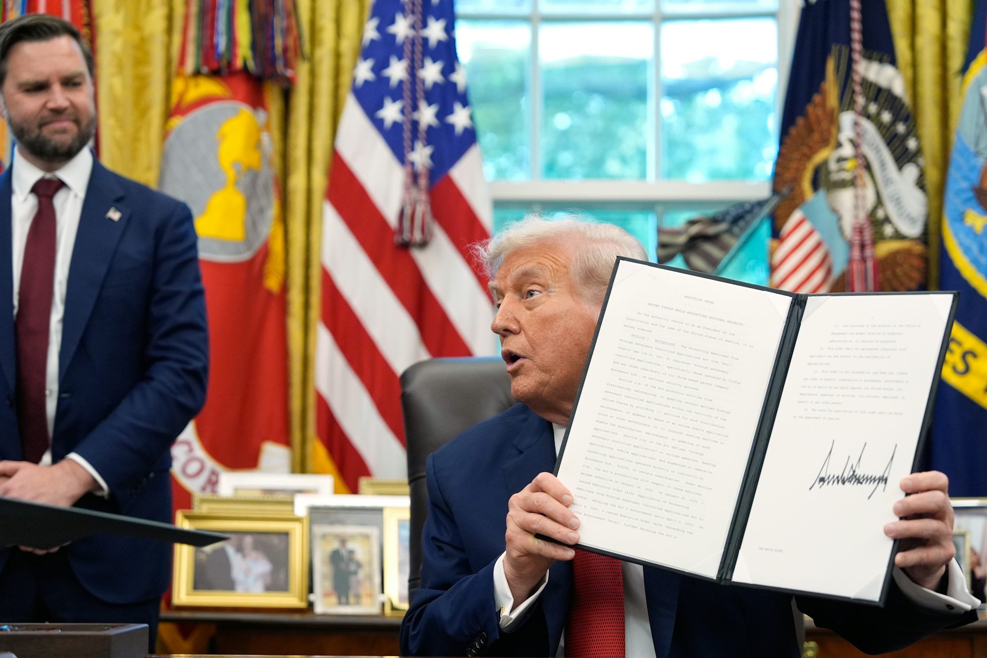 A man holds up a signed document, another man in a suit looks on, and there's an American flag in the background