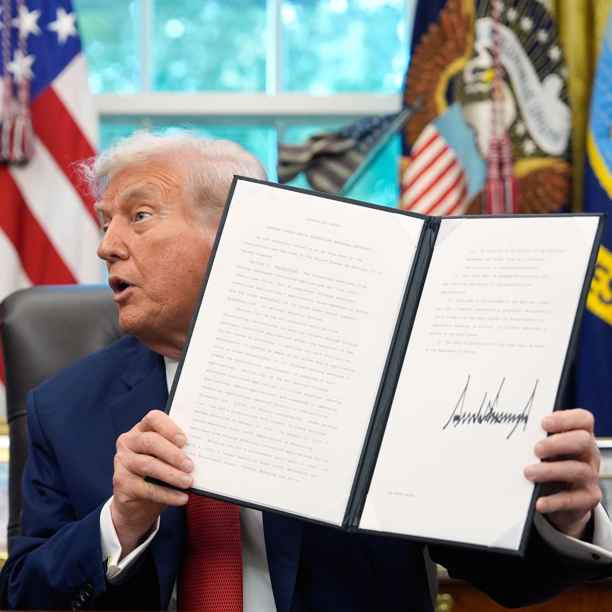 A man holds up a signed document, another man in a suit looks on, and there's an American flag in the background