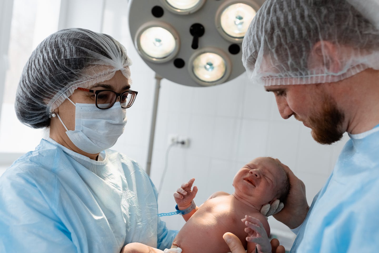 A man and woman, both dressed in protective gowns, hold up a newborn baby.