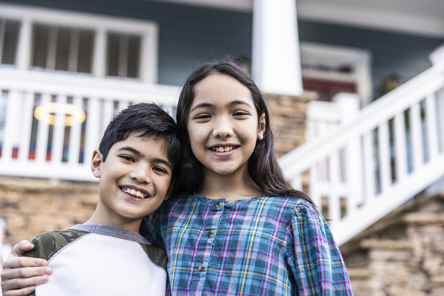 A young boy and older girl stand together smiling in front of a house.