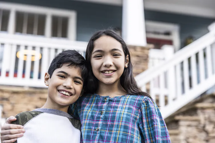 A young boy and older girl stand together smiling in front of a house.