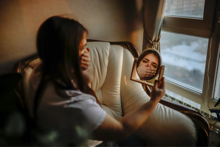 Girl covers her mouth while she looks in a mirror.