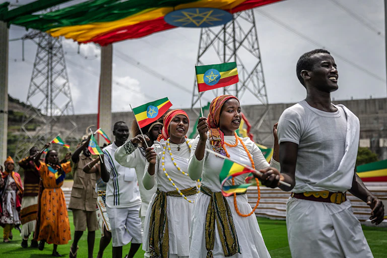 A line of smiling women and men dressed in white and holding small flags with green, yellow and red stripes