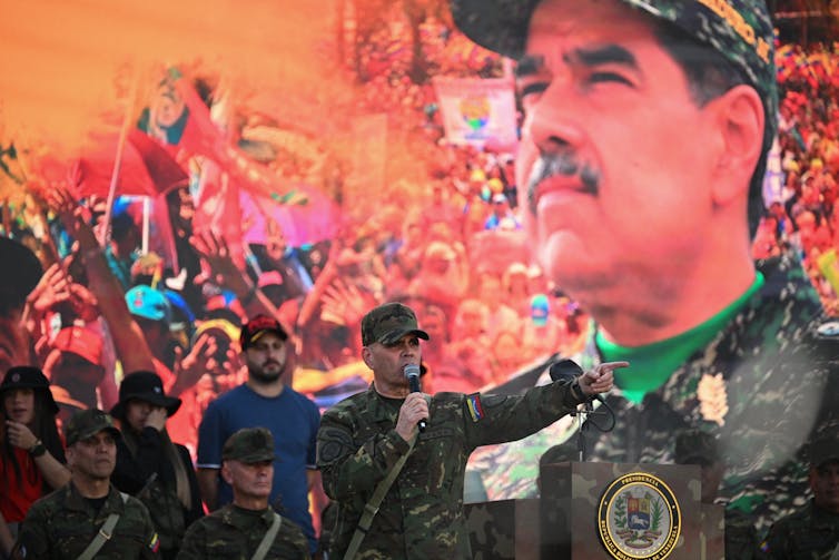 A man in army fatigues speaks and gestures in front of a large photo of another man.