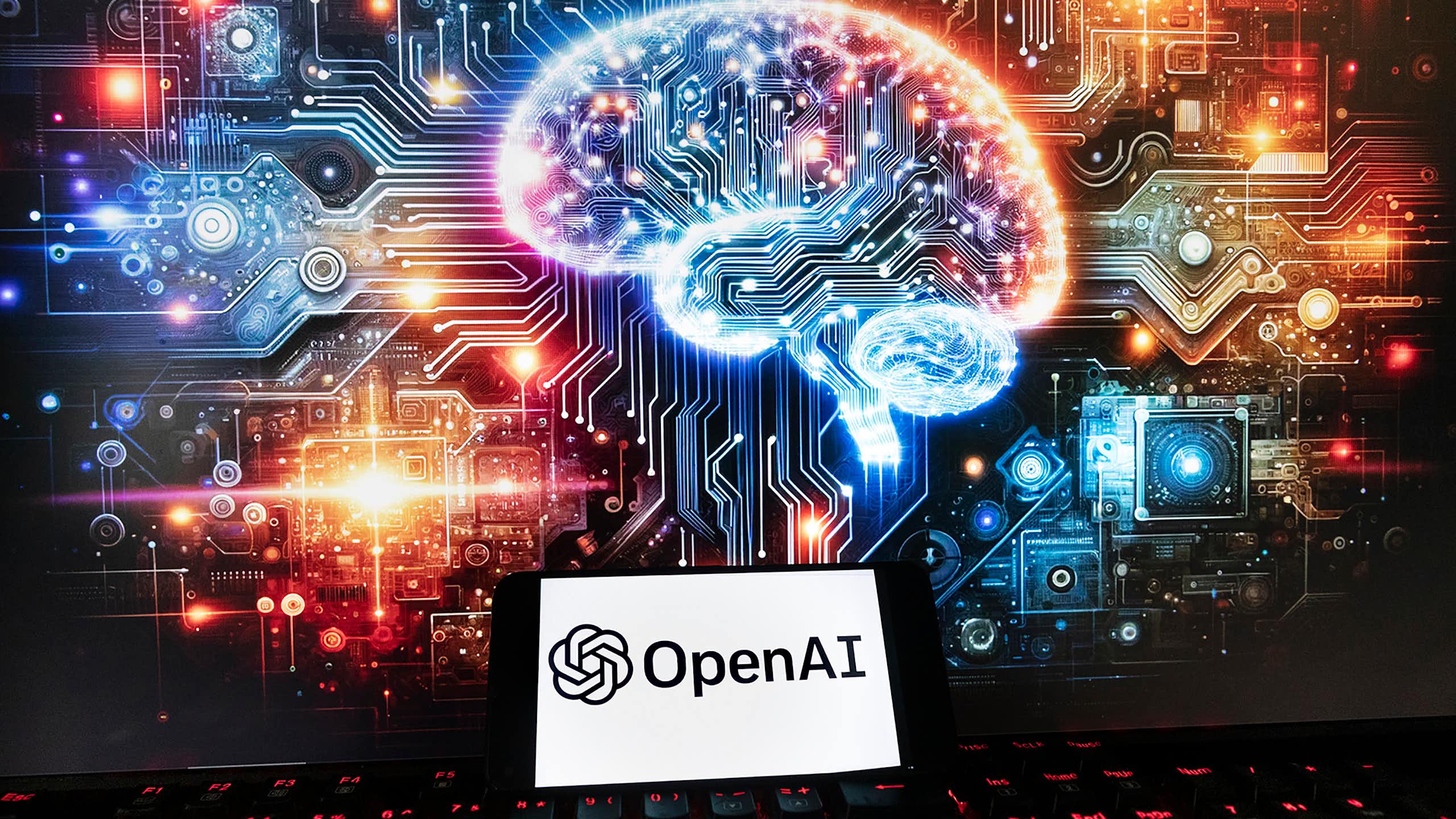 Le logo d’OpenAI est affiché sur un téléphone cellulaire, avec une image sur un écran d’ordinateur représentant un cerveau illuminé et entouré de lumières et de motifs multicolores.