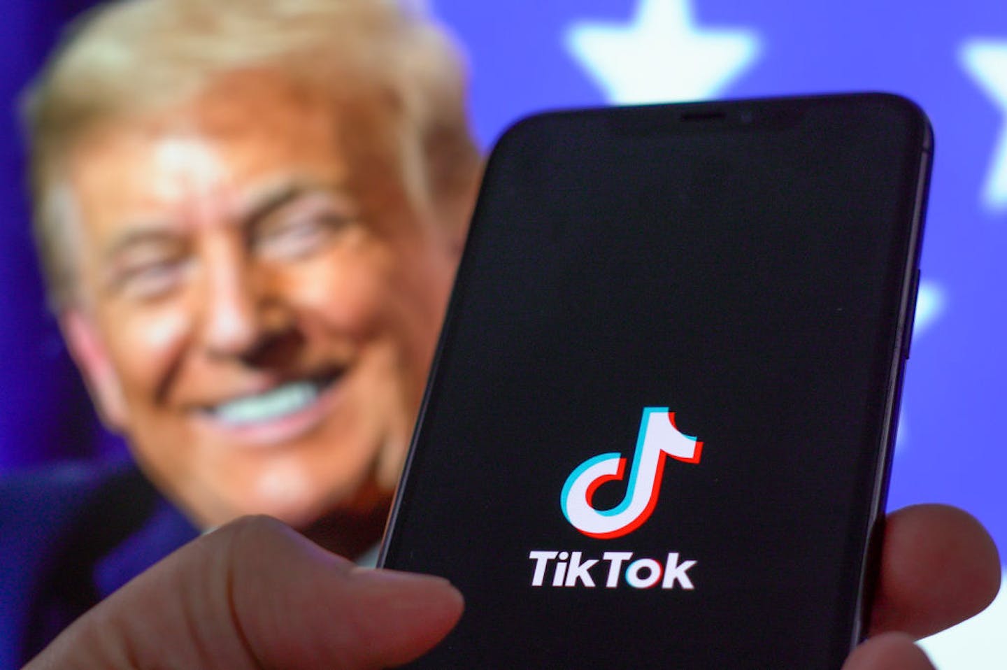 Hand holding mobile phone in foreground featuring the words 'TikTok,' while President Donald Trump smiles in the background.