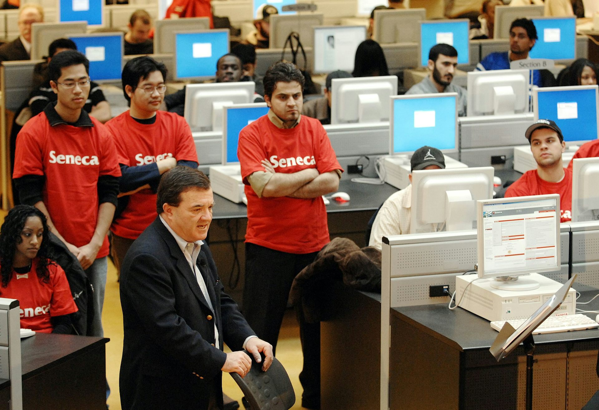 El hombre con el traje y los lazos en medio de un estudiante en la camiseta de Seneca y grandes monitores de computadora.