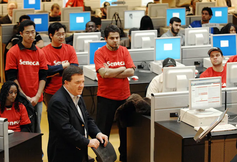 A man in a suit and tie amid student in Seneca t-shirts and large computer monitors.