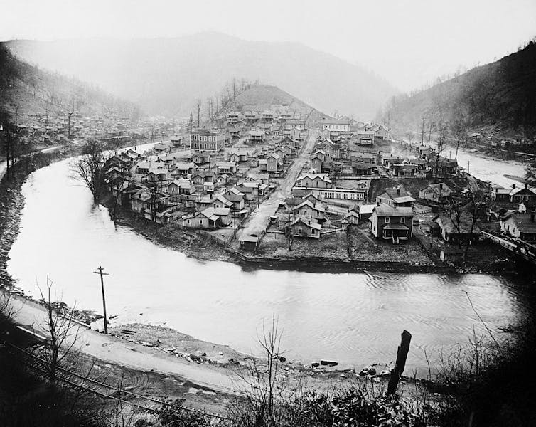 A black and white photo of a settlement on a small hill along a river.