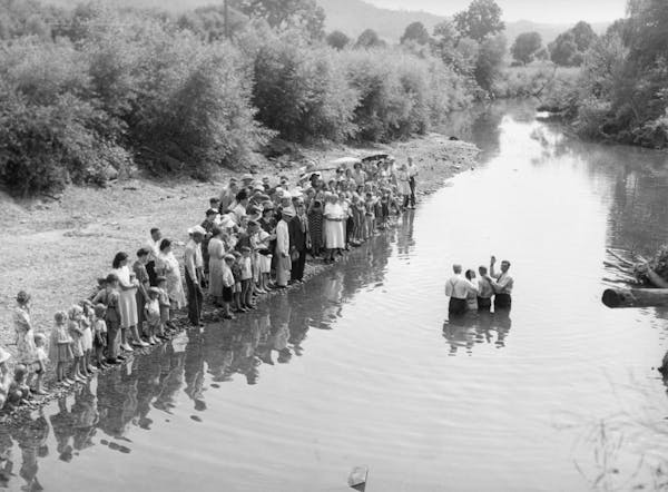 A black and white photo of a small crowd gathered along a riverbank as a few people stand in the middle, waist-deep.